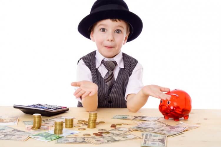 Sly boy with suit and hat at table with stacks of coins, bills, a piggy bank and a calculator. to illustrate how children and money matter.