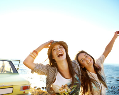 Two young women smiling with arms raised having the time of their lives at beach with car in background