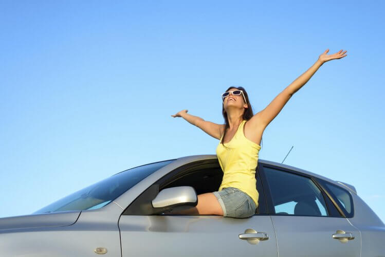 Joyful young driver sitting on car raising arms up to the sky on summer trip.