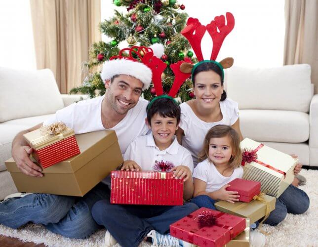 Happy young family of 4 celebrating Christmas at home with presents. Father wearing Christmas hat, mom and boy wearing reindeer ears.