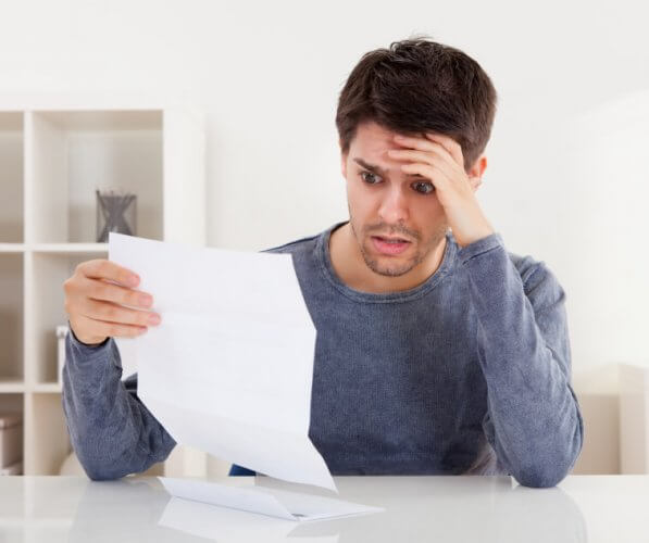 Horrified young man reading wide-eyed a document with an aghast expression and his hand to his forehead.