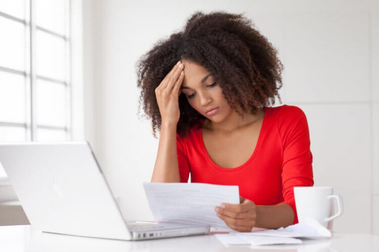 Young Black woman looking at papers next to her laptop and a cup of coffee. She looks concerned and holds hand on her temple.