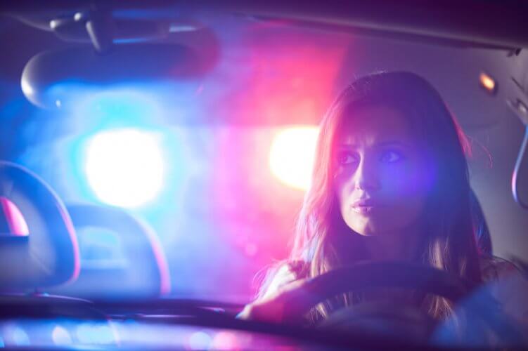 Concerned young female driver looking at the rear-view mirror. The lights of a police vehicle are seen in the background