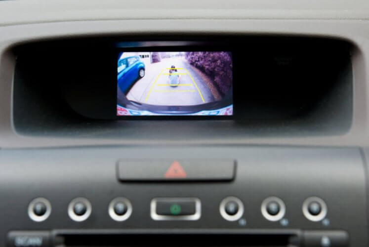 Close up to car dashboard with backup camera showing two kids sitting behind the car to illustrate the importance of having a backup camera in cars.