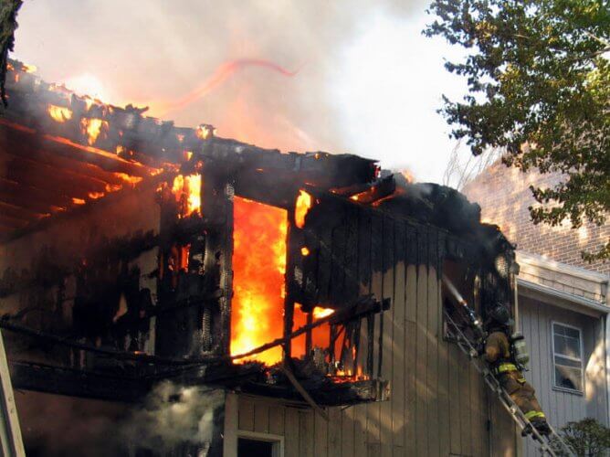 Top floor of a house on fire with fireman with water hose on ladder illustrating renters insurance coverage.