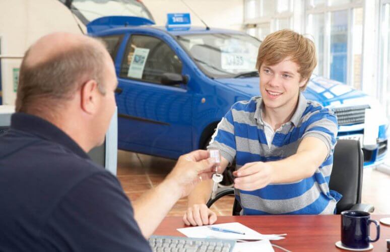 Older salesman handing the keys of a new car to a young man who just bought the car in the background to illustrate what is the best time to buy a car