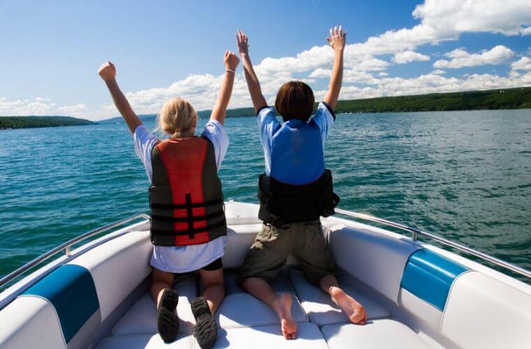 Rear view of couple kneeling on boat bow, wearing safety vests and raising their hands that illustrates 3 Types of Insurance You Need If You're Headed to the Water
