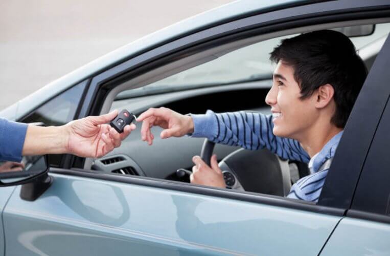 Teen driver on front seat of a car being handed the keys by an adult to illustrate whether it is time to re-examine the minimum driving age for teen drivers.