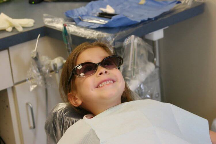 Child with Dental Health Insurance wearing protection sunglasses on dentist chair with equipment behind.