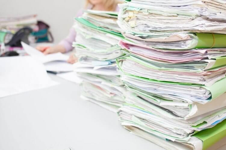 Photo of several stacks of medical files on desk and a woman behind them reading one to depict how Medi-Cal Backlog Leaves California Residents in Health Limbo