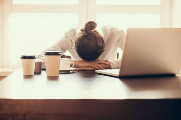 Tired woman resting her head on her arms over desk with 4 cups of coffee and a laptop.