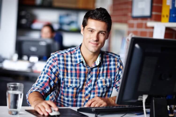 Smiling young man working on a desktop computer another worker behind him.