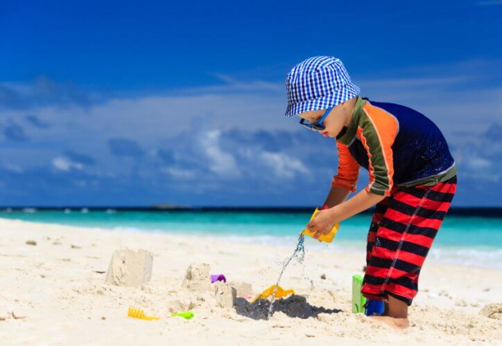 Kid at the beach playing with sand. He wears a one piece pinstripe swimsuit, hat and sunglasses.