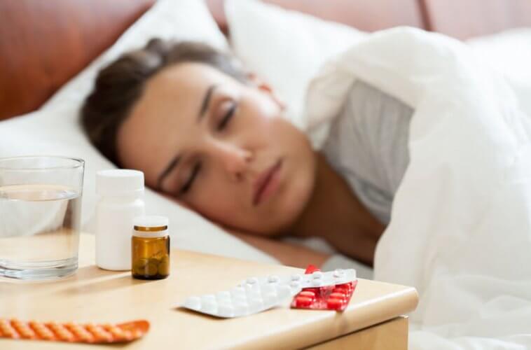 Nightstand with many pill cases next to a glass of water. Next to it, a young woman sleeping in bed