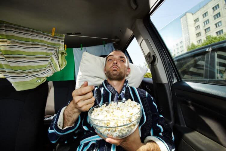 Man in car wearing bathrobe, laying his head on a pillow over the seat eating popcorn with a drying rack with clothes behind him to illustrate living in a car