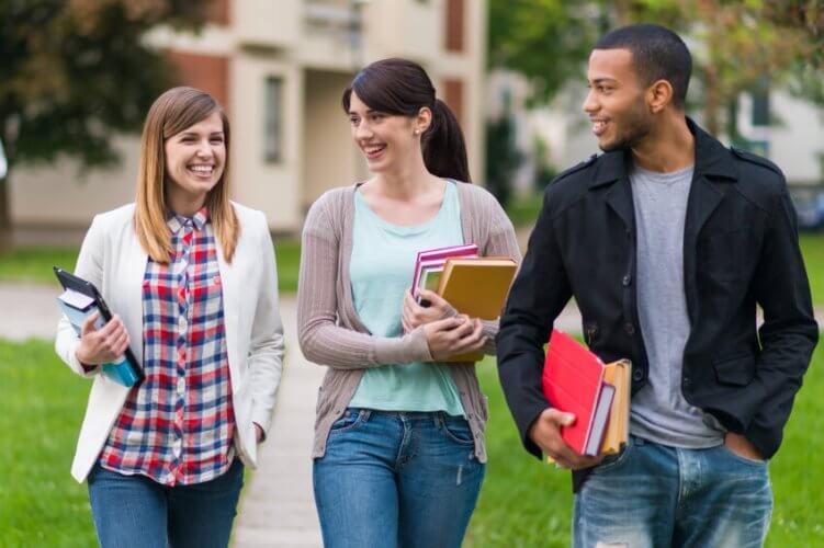Two young Caucasian women and one young Black man walking on campus holding books.