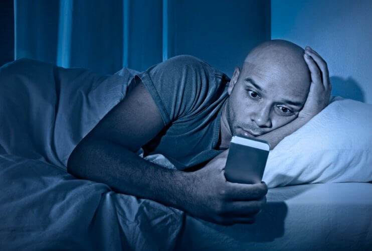 Young man awake at night in bed using smartphone because he is addicted to the internet