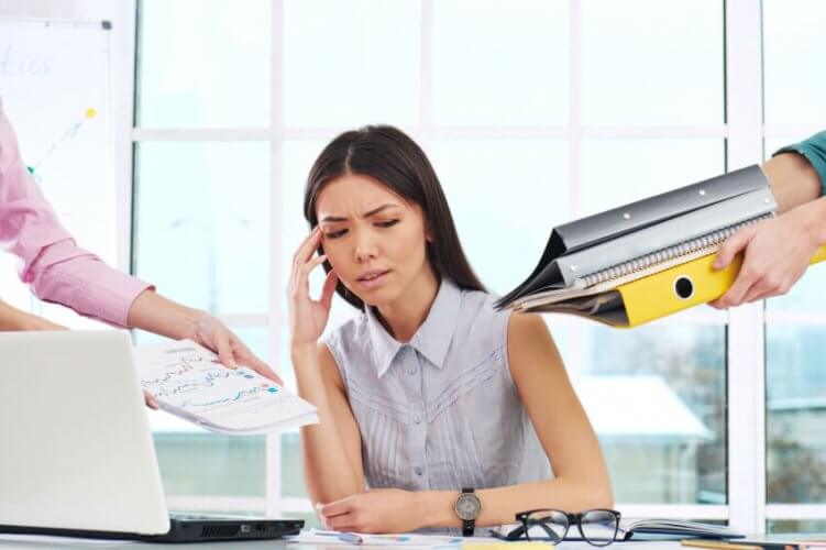 Young businesswoman stressed with hand on temple while two people hand her more documents to work on.