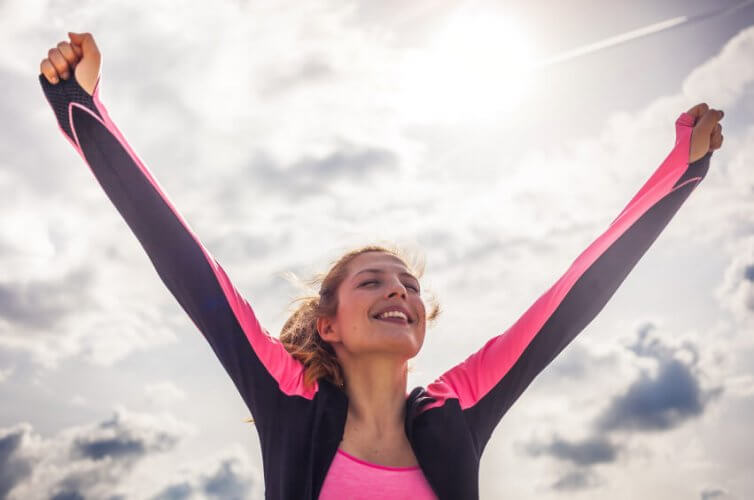 Happy young woman with her arms up in joy with a cloudy sky and the sun in background