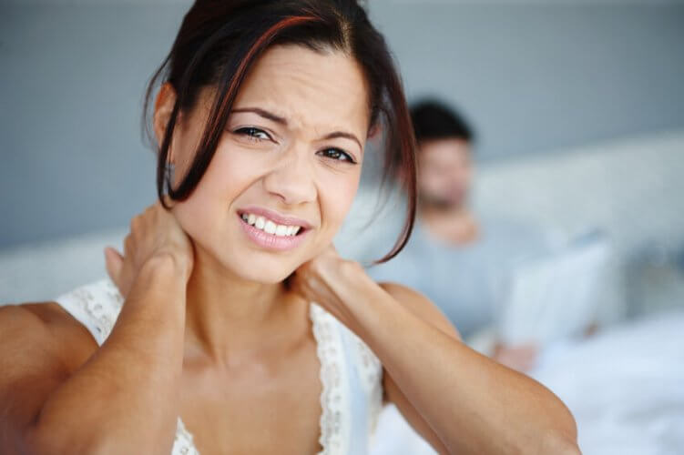 Close up of young woman with bad neck pain. Both hands holding back of her neck. On blurred background, her partner sitting on bed.