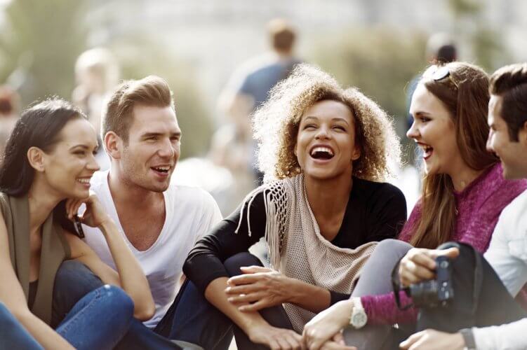 Multi-ethnic group of young adults sitting on grass chatting and laughing.