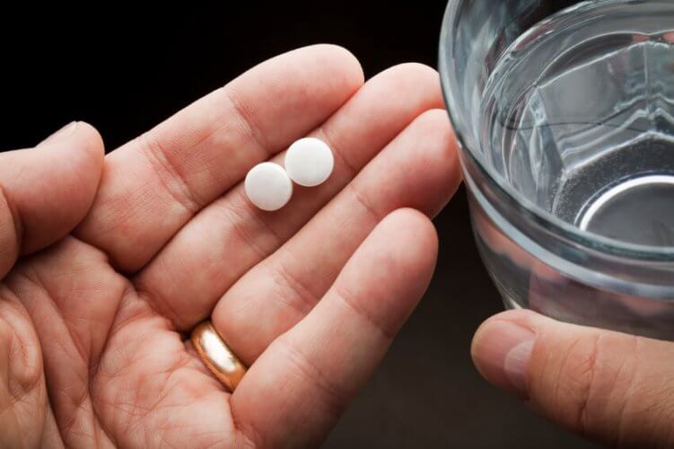 Close up to a pair of hands. Left hand with wedding ring holding two round pills and right hand holding a glass of water.