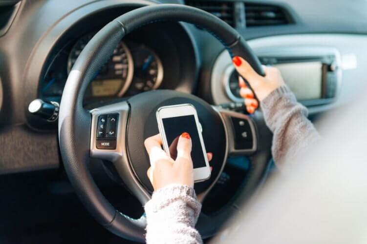 Picture of driver's hands texting and driving. Focus on car wheel and the smartphone over it held by the driver with one hand.