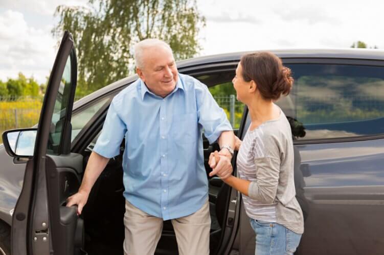 Smiling senior male driver getting out of the car with the aid of his daughter.