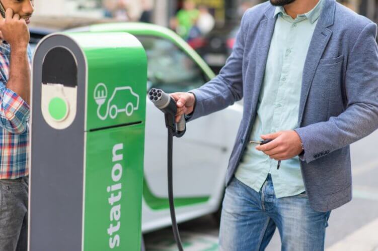 Electric car charging station. One young man puts back charging device. Another young man on the phone waits.