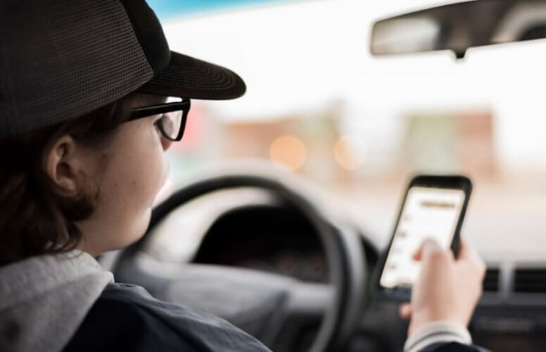 Rear view close up to a young driver wearing glasses and a cap, texting and driving.