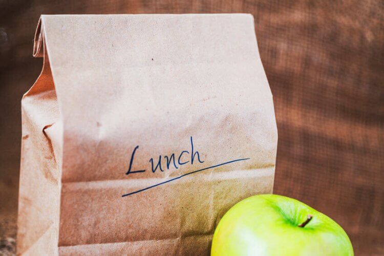 A paper bag that reads lunch next to an apple to illustrate How Bringing Your Own Lunch Can Help You Save Money