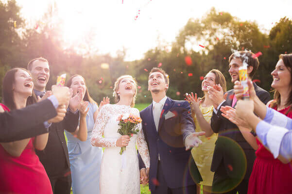 Happily married young couple surrounded by friends in wedding party in green sunny park showered with confetti