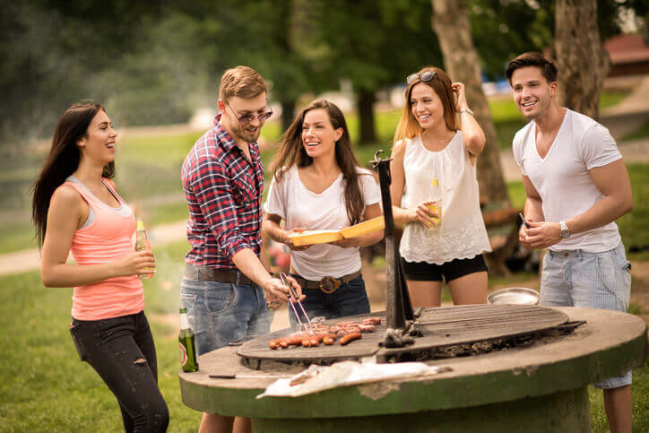 Young people (2 men, 3 women) having fun during barbecue picnic in park. One is cooking sausages. The others hold a beer and smile/chat.