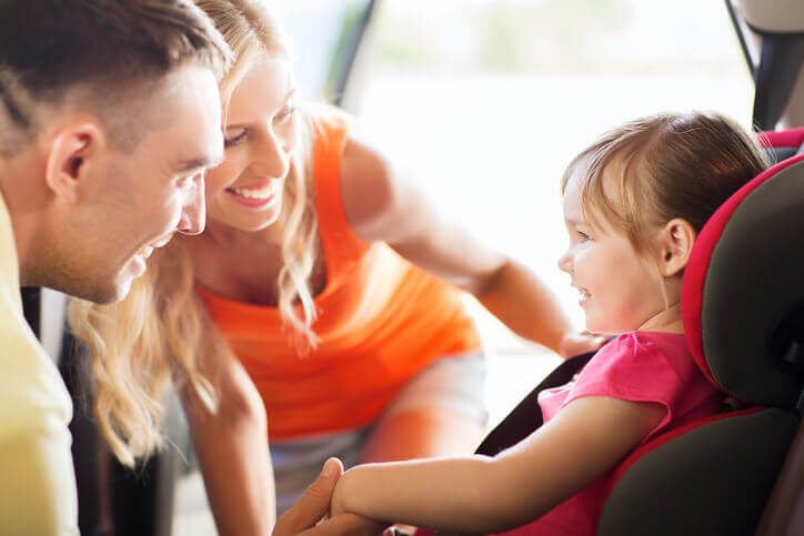 Happy young parents talking to smiling toddler girl in baby car seat to illustrate life insurance
