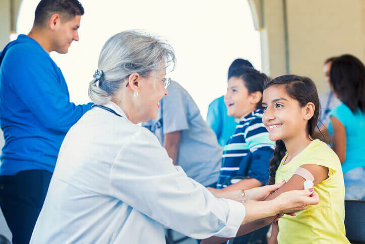 Smiling young Hispanic girl getting a bandaid from a senior female doctor after receiving her flu shot. Other volunteers and children behind them.