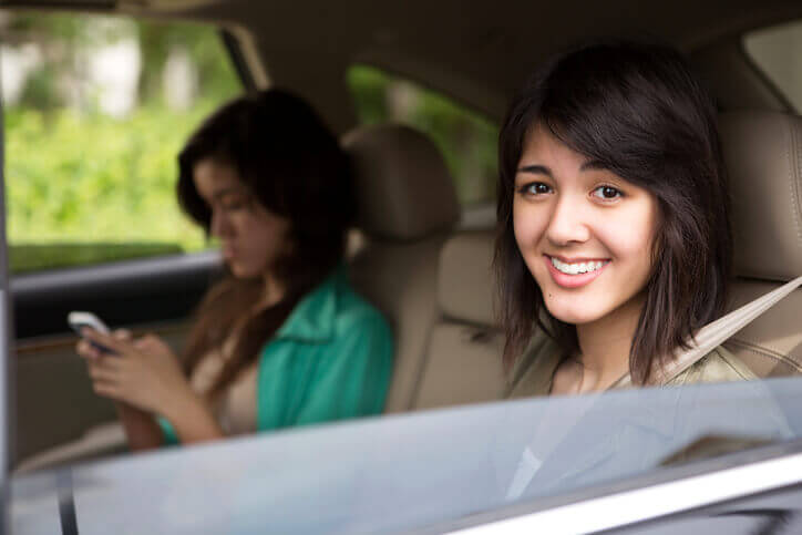 Female Hispanic teenager smiling to the camera from driver's seat. Female friend texting in passenger's seat. Both wear seatbelts.