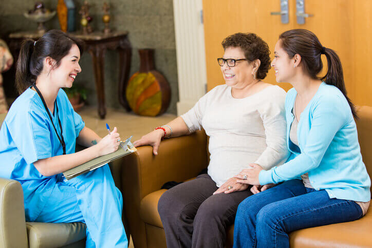 Nurse taking notes talking to Hispanic senior female patient who sits next to young Hispanic woman holding hands.