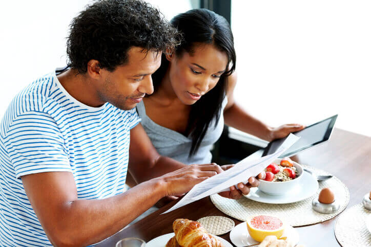 Young couple calculating home expenses holding papers and a tablet at breakfast table.