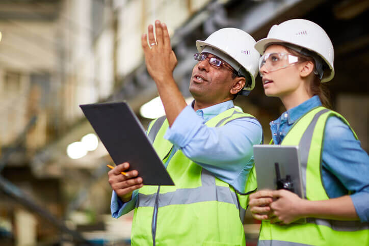 Male Hispanic senior engineer showing building to younger, female Caucasian engineer. Both wear helmets and security glasses.