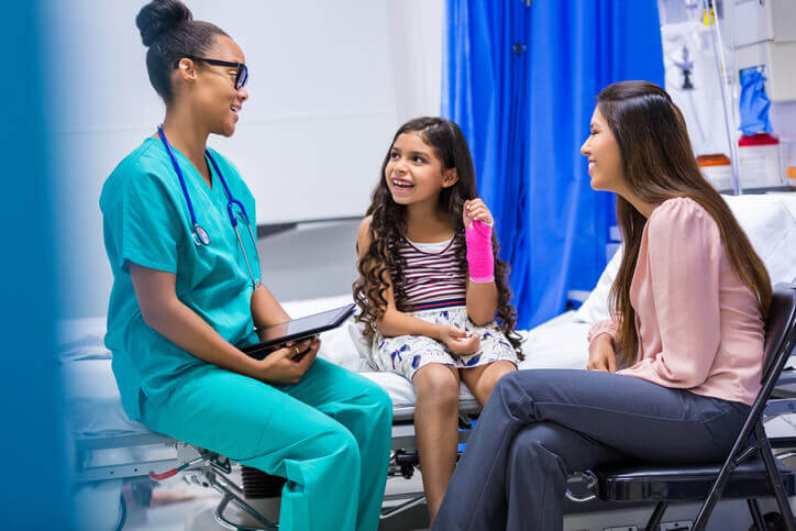 Young female Black nurse treating smiling Latin girl patient with broken arm in emergency room. Patient's sister sits next to her.