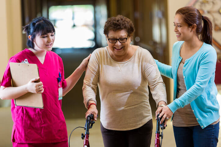 Happy Hispanic senior woman uses a walker in nursing home helped by young female physical therapist and the woman's granddaughter.