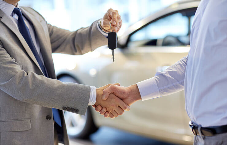 Close up of sales agent giving key to new car owner and shaking hands in auto showroom