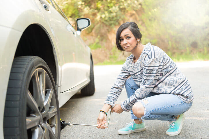 A young hispanic woman with a resigned face is stranded on the side of the road changing a flat tire.