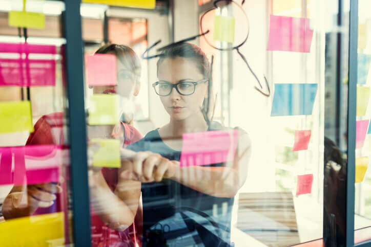 Two young businesswomen working together on wall glass with post it stickers.