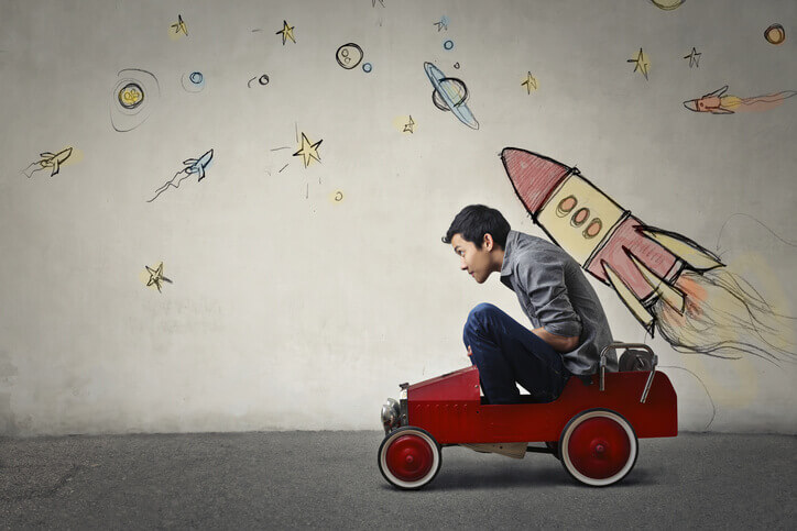 Young Asian guy in toy car in front of wall with spaceship and planet drawings. Spaceship seems to be attached to his back