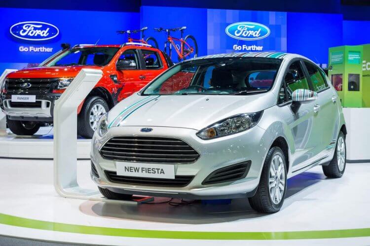 Ford Focus displayed at a Ford showroom. Behind it, a Ford Ranger pickup truck. Banners with Ford logo as background.