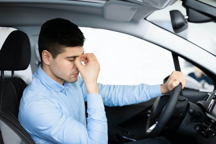 Tired young male driver with left hand on wheel and right hand on nose bridge.