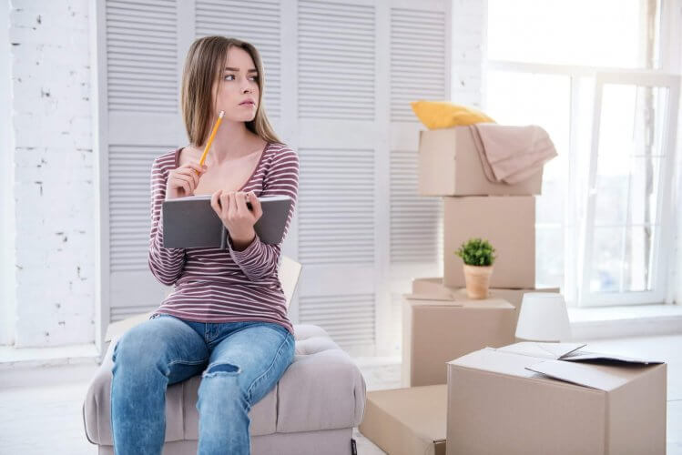 Young woman sitting on small couch in new apartment holding notebook and pencil. Moving boxes around her.