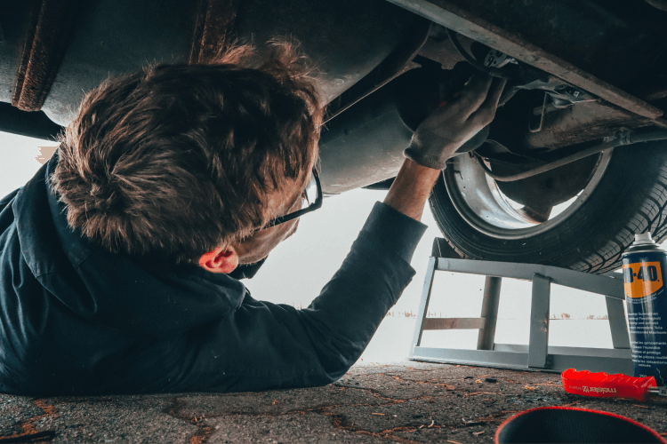 Young male mechanic working on car maintenance. He wears glasses and gloves and is lying on floor under car.