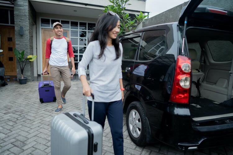 Young, happy, multi ethnic couple walking out of the house to place their luggage in the trunk of their van.